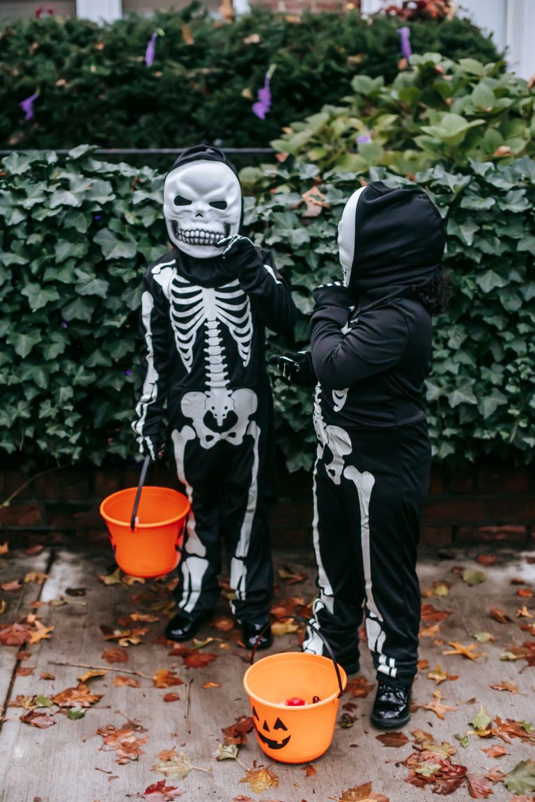 Two kids in skeleton costumes holding jack-o’-lantern candy buckets